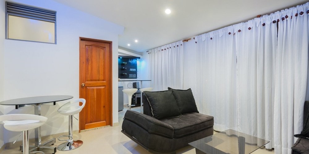 Sala de estar de un apartahotel con sofá oscuro, mesa de centro de vidrio y cortinas blancas hasta el techo