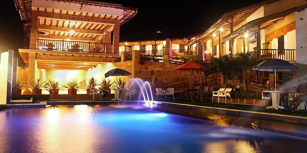Piscina de hotel colonial iluminada de noche con fuentes de agua, sombrillas y mesas junto a las habitaciones con balcón