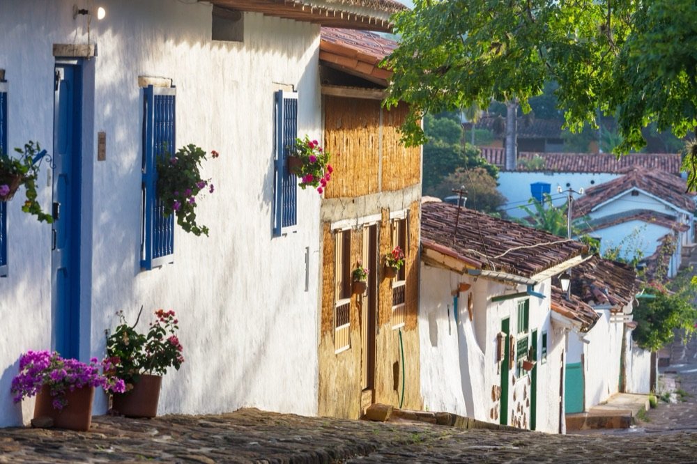 Calle empedrada de Barichara con casas de tapia pisada, puertas y ventanas azules, y macetas con flores moradas