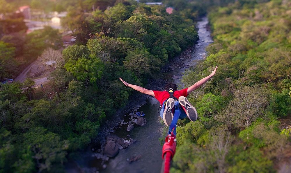 Vista aérea de una persona con camiseta roja saltando al vacío en bungee jumping sobre un río encajonado entre montañas boscosas