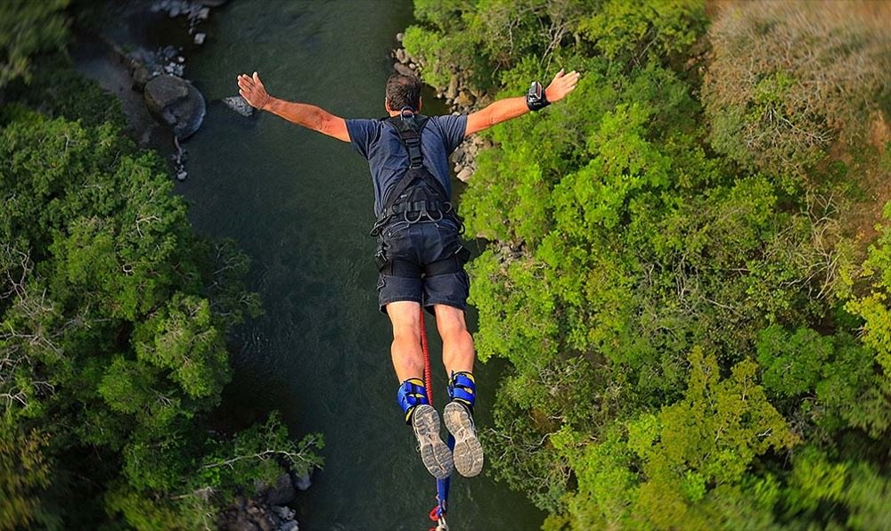 Hombre en caída libre de bungee jumping con los brazos abiertos sobre un río de aguas verdosas encajonado entre árboles