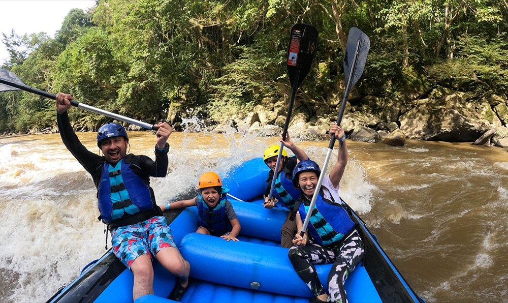 Familia con niños remando en una balsa azul mientras cruza un rápido del río Fonce, con las aguas color café salpicando