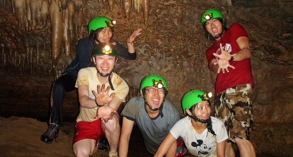 Grupo de exploradores con cascos verdes posando bajo estalactitas dentro de la Cueva de la Vaca, con las manos manchadas de barro