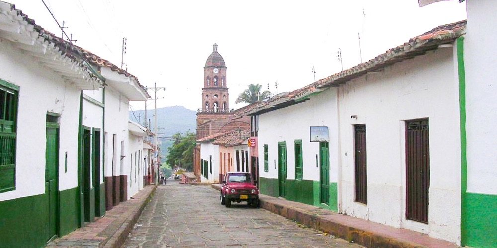 Calle empedrada de Curití con casas blancas de puertas verdes y un jeep rojo estacionado, con la torre de la iglesia al fondo