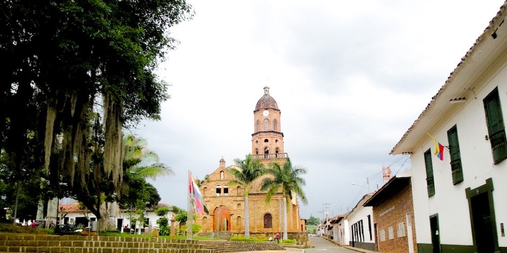 Fachada completa de la iglesia de Curití en piedra rojiza con su alto campanario, palmeras y la bandera de Colombia al frente