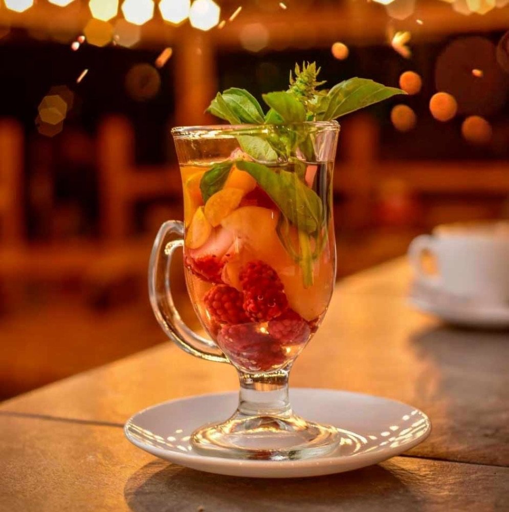 Copa de té caliente con frutos rojos, durazno y albahaca fresca sobre un plato blanco, iluminada por luces cálidas