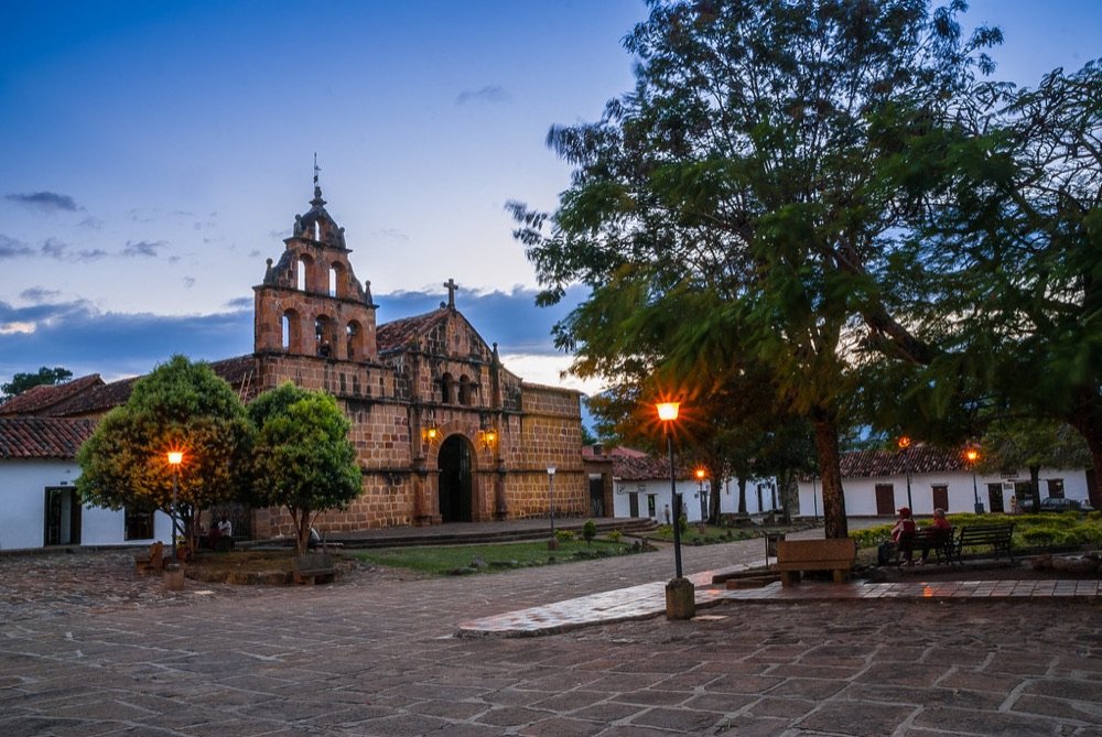 Fachada de piedra de la iglesia de Guane iluminada al atardecer, con la plaza y los faroles encendidos frente a ella