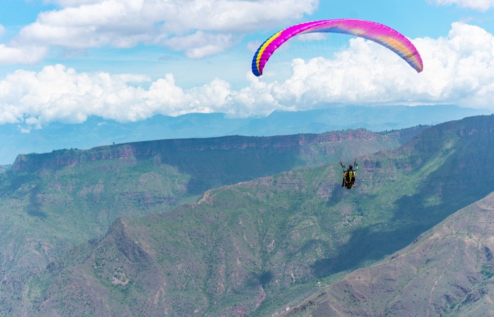 Parapente de vela rosada, azul y amarilla volando sobre las montañas del cañón del Chicamocha