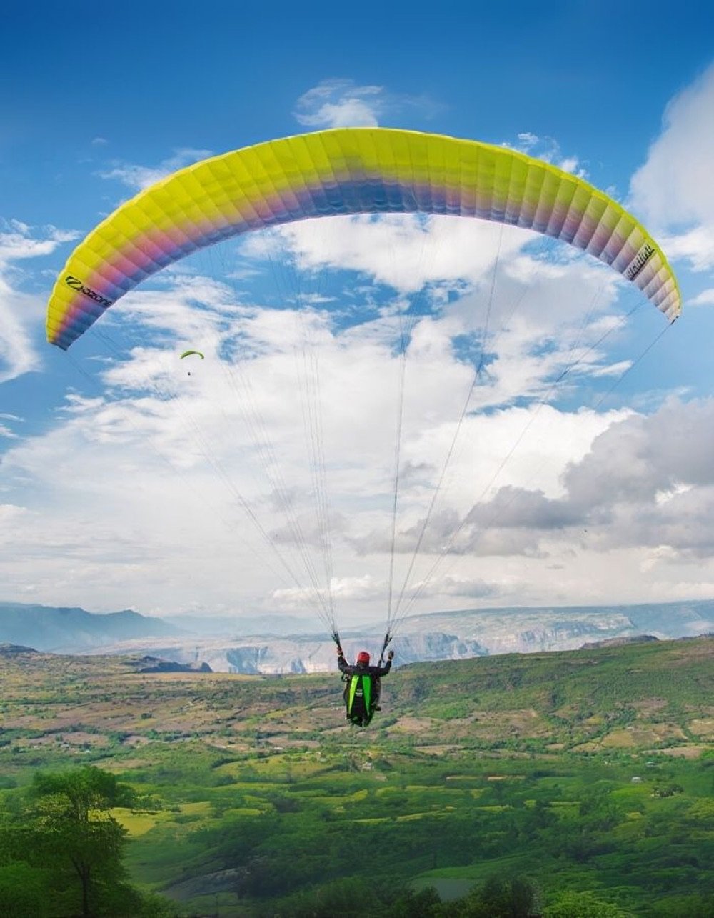 Parapente amarillo descendiendo sobre el valle verde cerca del voladero de Las Vueltas, en Curití, con las montañas de fondo
