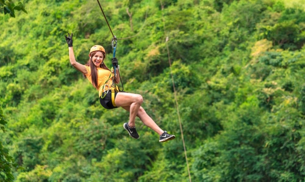 Mujer con casco naranja volando en tirolesa sobre el bosque, con las piernas extendidas y una mano en alto celebrando