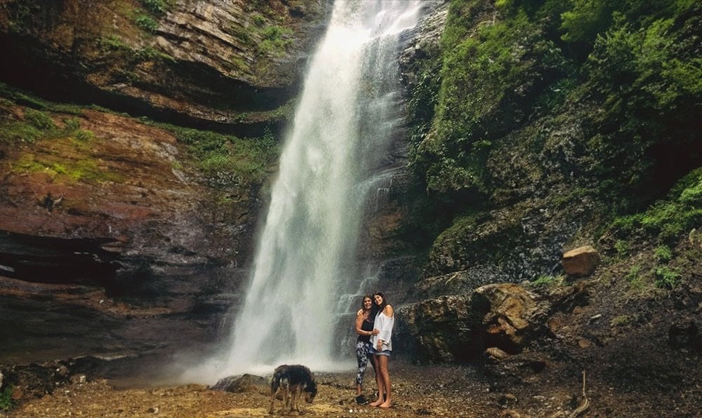 Dos mujeres y un perro posando al pie de la cascada de Juan Curí, con la caída de agua completa detrás de ellas