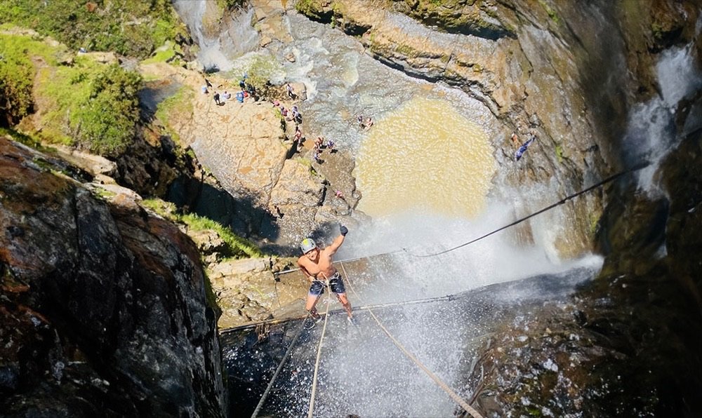 Vista desde arriba de un hombre descendiendo en rappel la cascada de Juan Curí, con turistas bañándose en la poza al fondo
